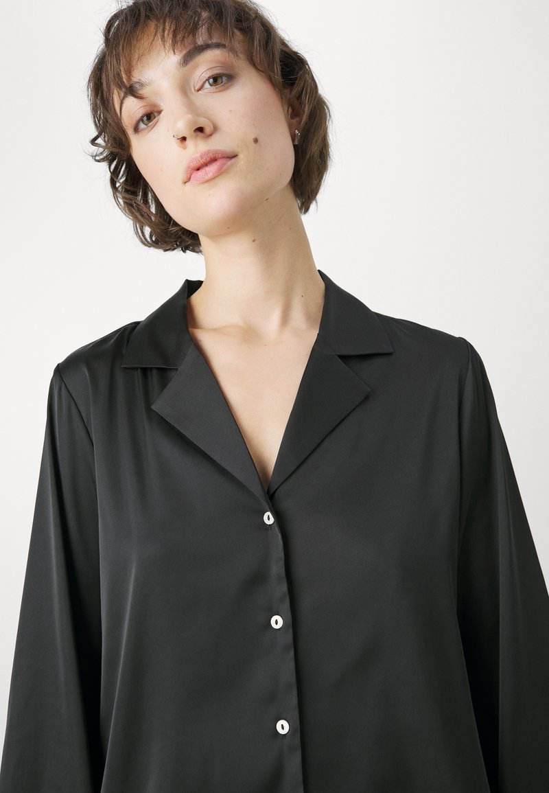 Camisa de satén negra con cuello clásico y mangas largas, que presenta un frente de botones con botones blancos y una textura suave.