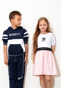 Felpa a strisce blu e bianche con logo "BUGATTI", abbinata a pantaloni coordinati. Ragazza in top bianco e gonna rosa con vita nera, presenta anch'essa il logo "BUGATTI".