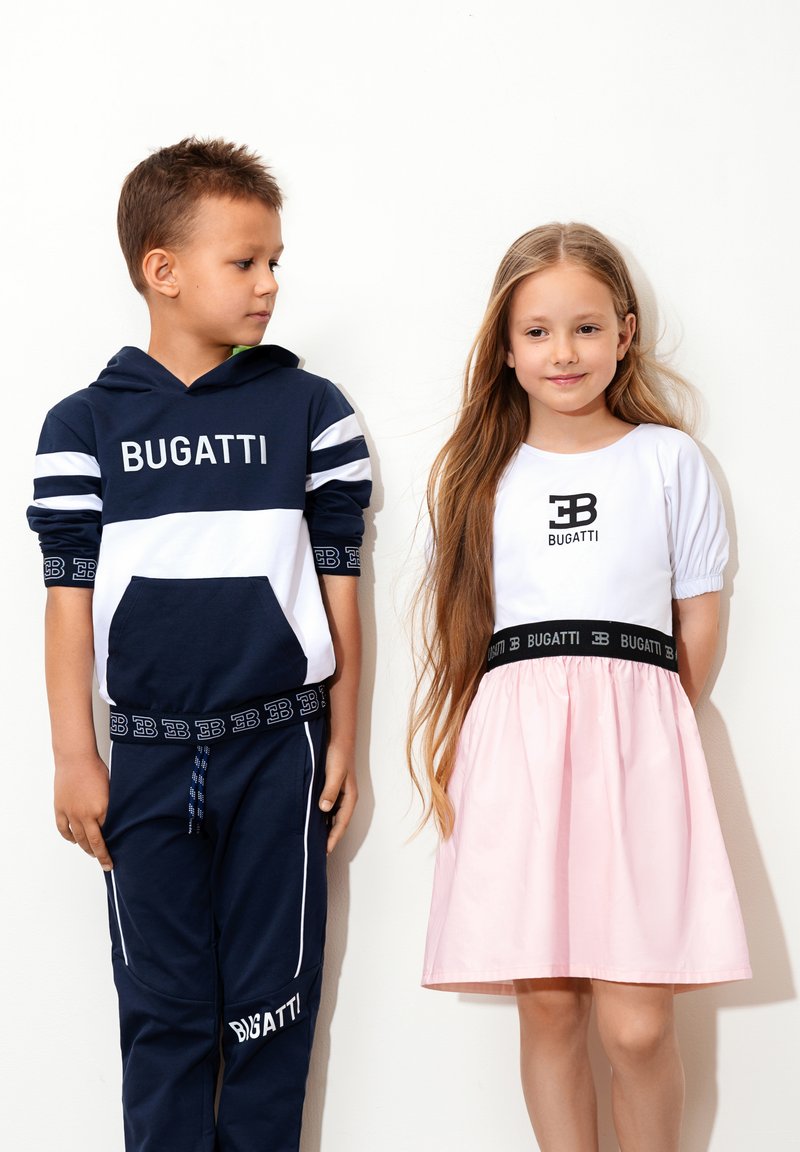 Felpa a strisce blu e bianche con logo "BUGATTI", abbinata a pantaloni coordinati. Ragazza in top bianco e gonna rosa con vita nera, presenta anch'essa il logo "BUGATTI".
