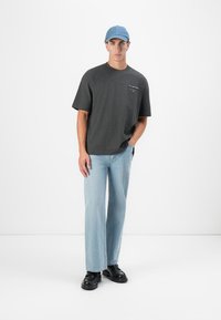 T-shirt gris foncé à manches courtes avec un petit logo blanc sur la poitrine, associé à un jean droit bleu clair et des chaussures noires. Casquette bleue.