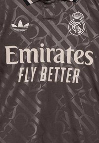 Maillot de sport gris foncé avec un motif subtil, un col polo, et le texte "Emirates FLY BETTER." Les logos d'Adidas et d'un club de football sont visibles.
