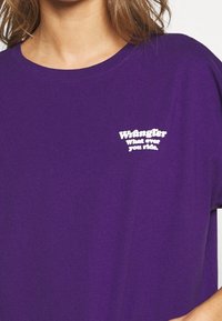 Lila bomullst-shirt med korta ärmar och rund halsringning. Framstycket visar "Wrangler"-logotypen tillsammans med frasen "what ever you ride."