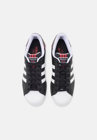 Czarne skórzane sneakersy z trzema białymi paskami, białą gumową osłoną na palce i czarnymi sznurówkami. Czerwono-biała marka na języku.