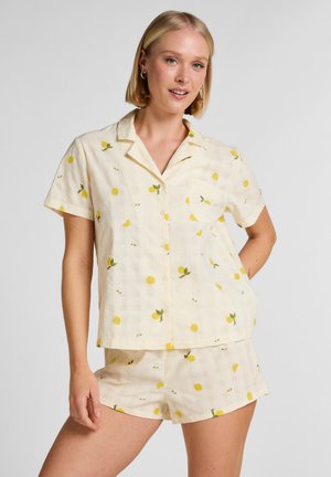 Femeie purtând un tricou cu nasturi cu mâneci scurte și pantaloni scurți asortați, cu imprimeu galben de lămâie pe material deschis, stând cu o mână pe șold.