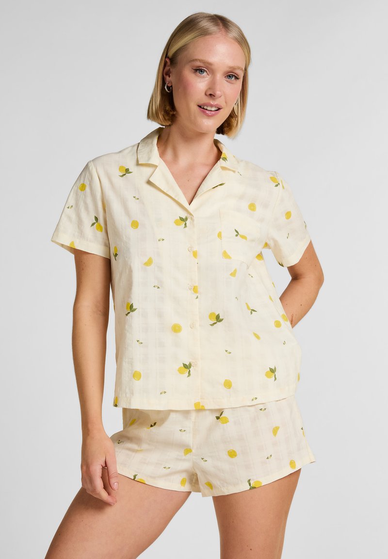Γυναίκα που φοράει ταιριαστό πουκάμισο με κουμπιά και κοντά μανίκια καθώς και σορτς με κίτρινο σχέδιο λεμονιών σε ανοιχτό ύφασμα, στέκεται με το ένα χέρι στη μέση.