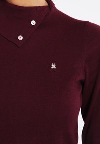 Pull en tricot bordeaux avec col, doté de deux boutons blancs et d'un petit logo brodé sur la poitrine. Texture lisse, coupe ajustée.