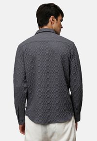 Camisa de manga larga texturizada de color gris con un patrón geométrico, que presenta un cuello y puños abotonados. La tela parece ligera y con diseño.