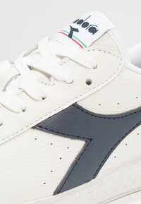 Sneaker in pelle bianca con logo e dettagli in blu navy scuro, superficie testurizzata, lacci bianchi e linguetta con logo.
