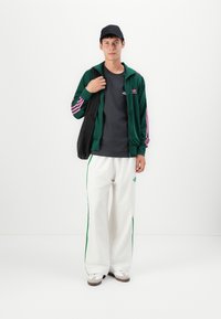 Grønn velour zip-up jakke med rosa striper, svart t-skjorte, hvite bukser med grønne sidestropper, bærende en svart tote bag, iført en svart caps.