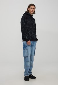Felpa nera con motivo marmorizzato, tasca a marsupio e cappuccio regolabile. Jeans cargo blu con molteplici tasche. Sneakers nere.