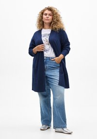 Cardigan bleu marine à ouverture sur le devant, avec des poignets côtelés et deux poches frontales, porté par-dessus un t-shirt graphique blanc et un jean taille large bleu clair.