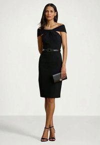 Γυναίκα φορώντας ένα εφαρμοστό μαύρο φόρεμα χωρίς ώμους με ζώνη, κρατώντας μια μαύρη τσάντα και φορώντας μαύρα παπούτσια με λουράκια στους αστραγάλους σε απλό φόντο.