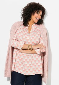Camicia leggera con base crema e motivi geometrici corallo, abbinata a un morbido cardigan aperto rosa, indossata sopra jeans scuri. Braccialetti dorati completano l'outfit.