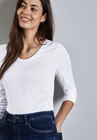 Weißes Langarmhemd mit rundem Ausschnitt, kombiniert mit dunkelblauen Jeans aus Denim mit schmaler Silhouette und sichtbaren Nähten.