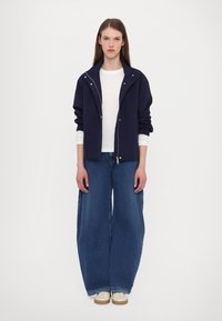 Navy blue jacket with a zip closure and large collar, paired with a white long-sleeve shirt and wide-leg denim jeans. Light-colored shoes.