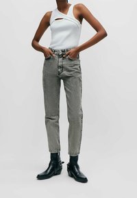 Valkoinen hihaton paita, jossa on aukollinen yksityiskohta, yhdistettynä harmaisiin korkeavyötäröisiin farkkuihin ja mustiin nilkkuriin. Kankaan rakenne on muotoiltu ja istuva.