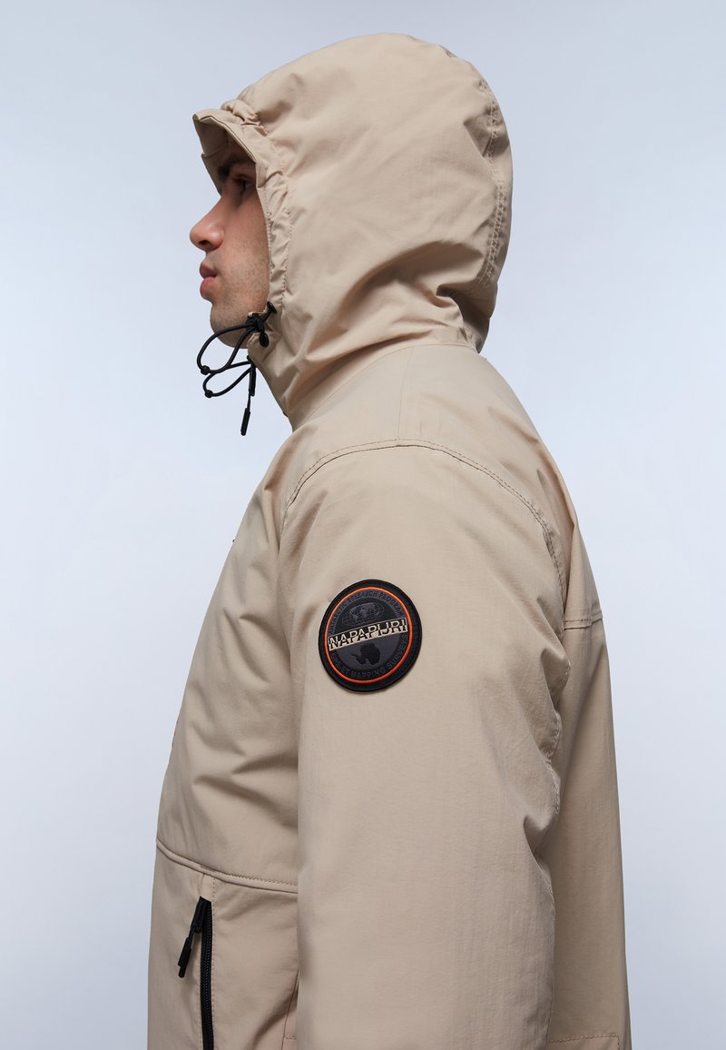 Beige gewatteerde jas met een gestructureerde afwerking, voorzien van een logopatch op de linker mouw en een ritszak aan de voorkant.