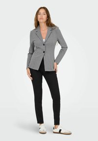 Femme debout aux longs cheveux, portant un blazer gris boutonné, un pantalon noir, un haut blanc et des baskets blanches à rayures noires.