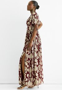 Maxi dress in burgundy fabric with gold floral patterns; features short puff sleeves and a thigh-high slit. Worn with strappy silver heels.