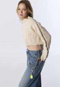 Jeune femme portant un pull en tricot torsadé de couleur crème, à manches courtes et détail de zip, avec un jean bleu orné de chaînes en argent superposées et de charms colorés.