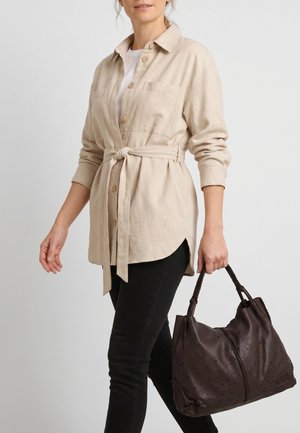 Vrouw draagt een beige blouse met ceintuur en zwarte broek, houdt een grote donkerbruine leren handtas met gevlochten handvatten vast.