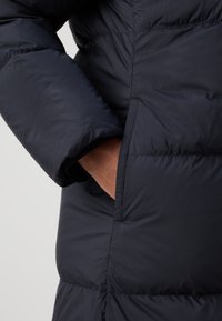 Die Hand einer Person steckt in der Seitentasche einer schwarzen, gepolsterten Winterjacke mit sichtbaren Nähten und einer glatten Stofftextur.