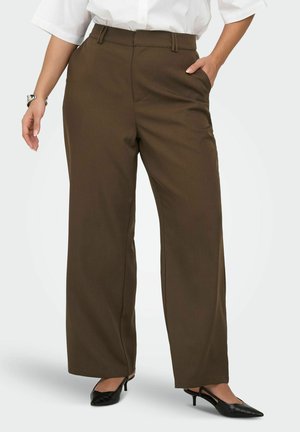 Mujer que lleva pantalones anchos de color marrón oscuro, camisa blanca metida, tacones negros de punta, con una mano en el bolsillo y reloj en la muñeca izquierda.