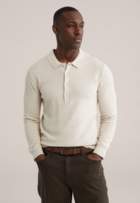 Polo à manches longues beige texturé avec un col polo et quatre boutons, associé à un pantalon marron foncé et une ceinture marron.