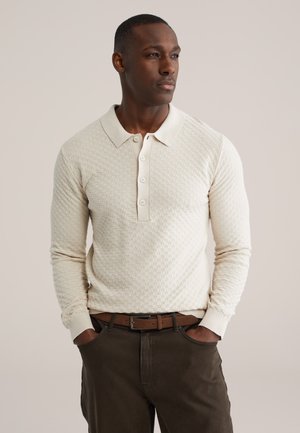 Beige getextureerd poloshirt met lange mouwen, een kraag en vier knopen, gecombineerd met donkerbruine broek en een bruine riem.