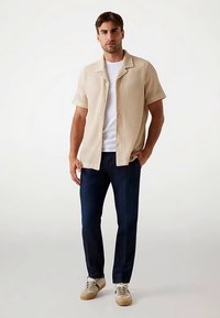 Beiges kurzärmeliges Rippshirt, weißes Unterhemd, blaue Jeans und beigefarbene Turnschuhe mit grünen Akzenten, stehend vor einem neutralen Hintergrund.