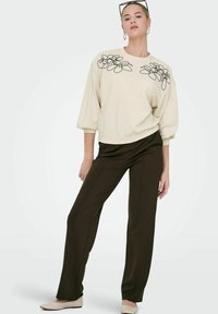 Krämfärgad sweatshirt med svart blommig broderi, breda ärmar, ihopparad med mörkbruna skräddarsydda byxor och beigea slip-on skor.