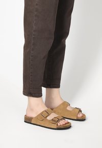 Brown suede sandals with two adjustable straps, cork footbed, and a rubber sole; worn with brown cuffed jeans.