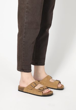 Brown suede sandals with two adjustable straps, cork footbed, and a rubber sole; worn with brown cuffed jeans.