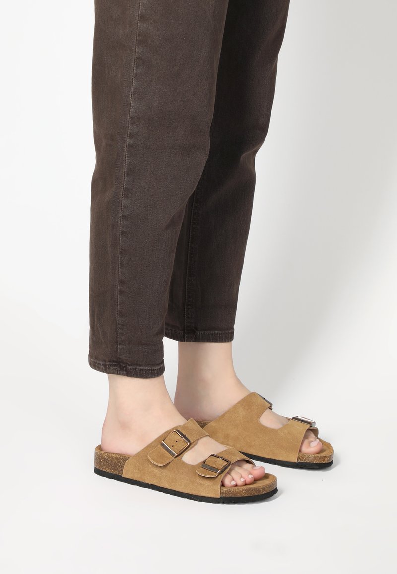 Brown suede sandals with two adjustable straps, cork footbed, and a rubber sole; worn with brown cuffed jeans.