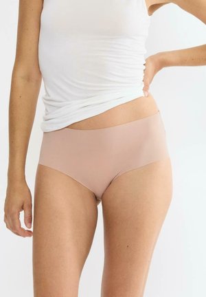 Kobieta w cielistych bezszwowych majtkach typu hipster i białym bezrękawniku, z ręką opartą na biodrze, na białym tle.