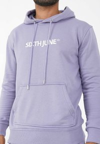 Sudadera de color púrpura claro con un bolsillo frontal, cordones ajustables y un logotipo en letras blancas en el pecho. Textura de tela suave.