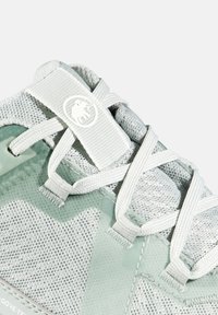 Lys grå atletisk sko med et mesh-overdel og teksturerede detaljer, flade hvide snørebånd og et logo-tag på tungen.