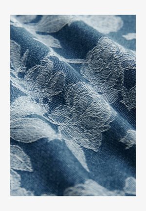 Denim tkanina z belim cvetnim vzorcem, teksturirana površina z globoko modro podlago, ki poudarja zapleteno zasnovo in material.