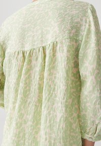 Blusa verde chiaro con un motivo testurizzato in rosa pallido. Presenta una parte posteriore arricciata e maniche a tre quarti con bottoni ai polsini.