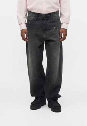 Personne portant un jean noir ample avec des éclaboussures de peinture blanche et des chaussures noires, associée à une chemise rose clair à manches longues.