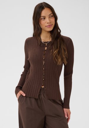 Tânără cu păr lung care poartă un cardigan maro striat, mulat, cu nasturi în față și pantaloni maro asortați, mâinile în buzunare, uitându-se pe lângă.