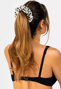 Wiązanka w wzór lamparta w białym i czarnym kolorze, trzymająca długie, proste brązowe włosy. W tle widoczne są czarne, teksturowane ramiączka od biustonosza.