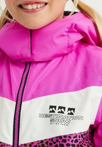 Barn som bär en rosa och vit snöjacka med leopardmönster och "Destination Snow"-logotyp på bröstet, som ler lätt.