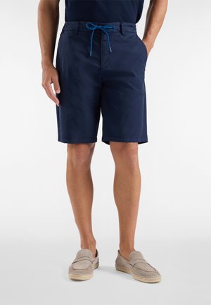 Uomo che indossa pantaloncini blu navy con coulisse e mocassini beige senza lacci, in piedi con una mano in tasca su sfondo bianco.