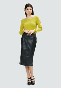 Maglione verde a maglia, design off-shoulder, abbinato a una gonna di pelle nera con dettaglio della fibbia e texture liscia. Scarpe con zeppa nere.