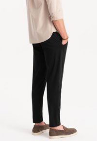Pantalones negros de corte relajado con bolsillos laterales; tejido suave y diseño de pierna entallada. Combinados con zapatos marrones sin cordones.
