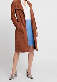 Manteau long en daim marron avec un col à revers, associé à une chemise claire et une jupe crayon bleue. Les chaussures présentent un motif imprimé serpent.