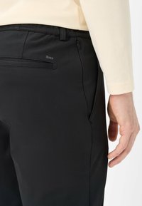 Pantalon de sport noir à texture lisse, avec une poche latérale zippée. Logo "Brax" visible à l'arrière.