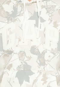 Felpa bianca con un motivo a foglia in grigio chiaro e beige, con un grande logo bianco "GAP" sul petto e un cappuccio regolabile con cordini.