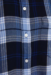 Gros plan sur une chemise à carreaux bleu et marine avec des boutons blancs et une poche poitrine visible sur le côté droit.